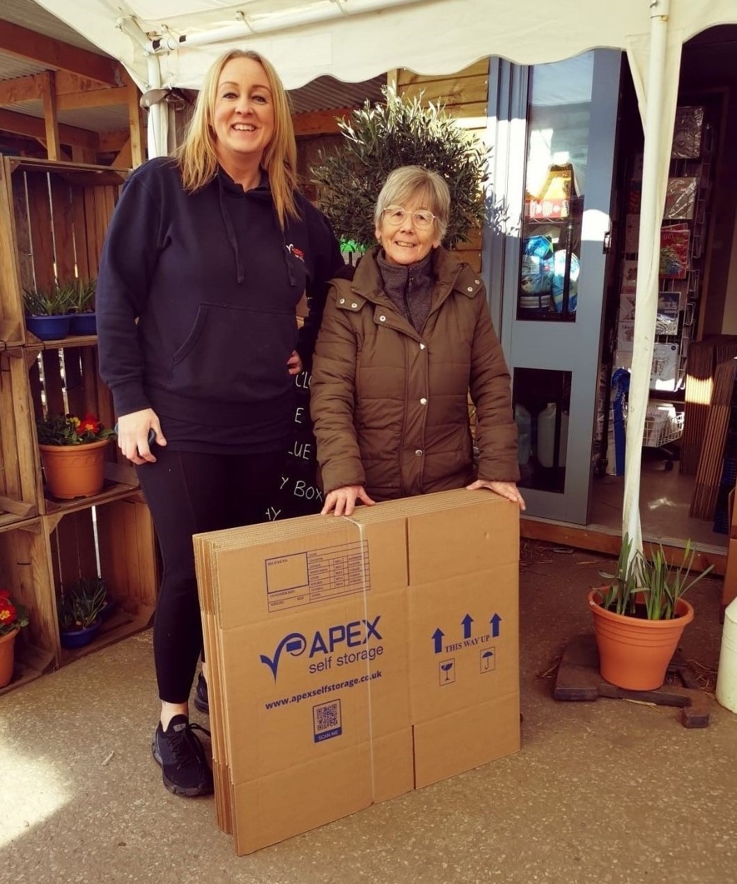 Apex team member smiling with volunteer from Biddulph Rotary Club, holding a stack of Apex Self Storage boxes